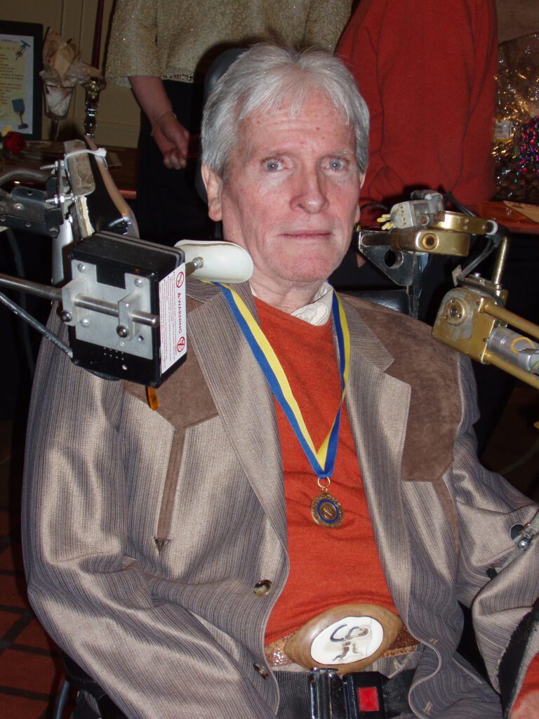 A man sits in a wheelchair with a command box by his face. He is wearing a red shirt with a brown, Western cut jacket, and medal around his neck.